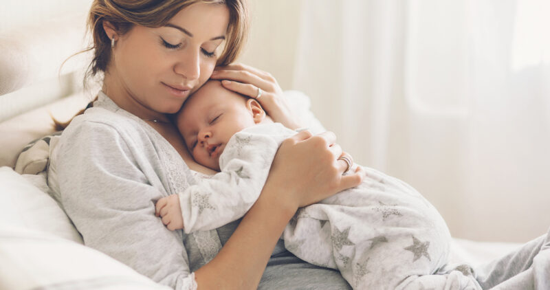Loving mom carying of her newborn baby at home. Bright portrait of happy mum holding sleeping infant child on hands. Mother hugging her little 2 months old son.